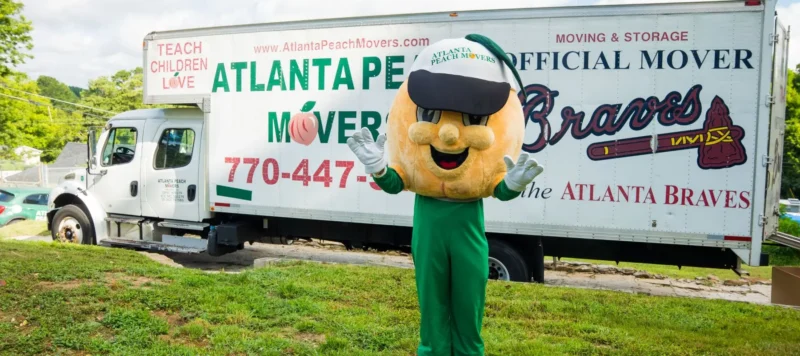 A person in a peach mascot costume stands waving in front of an Atlanta Peach Movers truck labeled as the official mover of the Atlanta Braves.