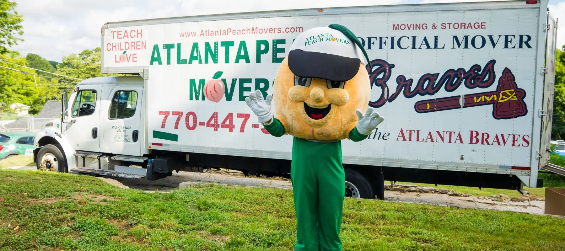 A person in a peach mascot costume stands waving in front of an Atlanta Peach Movers truck labeled as the official mover of the Atlanta Braves.