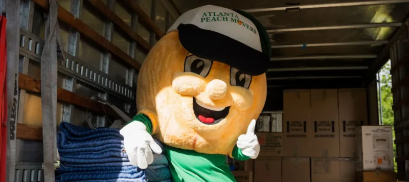 A person in an oversized peach mascot costume with a hat that reads Atlanta Peach Movers stands inside a moving truck with boxes and moving blankets.