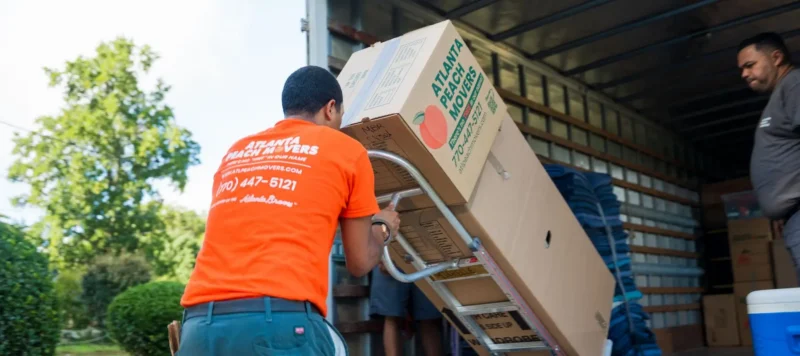 A mover in an orange shirt uses a dolly to load a large box onto a moving truck. Another mover is inside the truck, and trees are visible in the background.