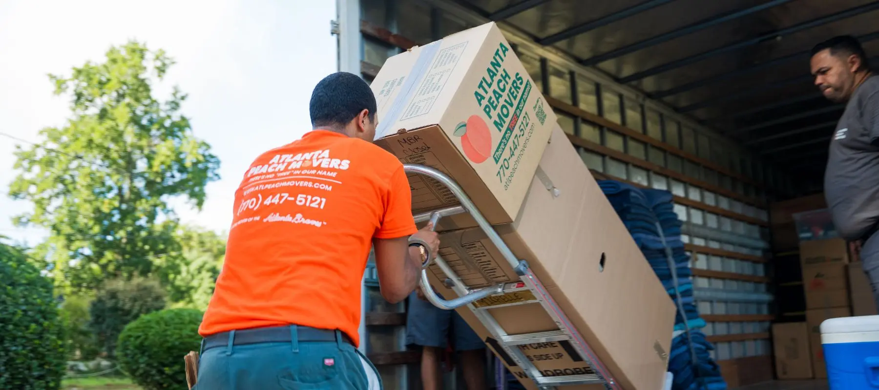 A mover in an orange shirt uses a dolly to load a large box onto a moving truck. Another mover is inside the truck, and trees are visible in the background.