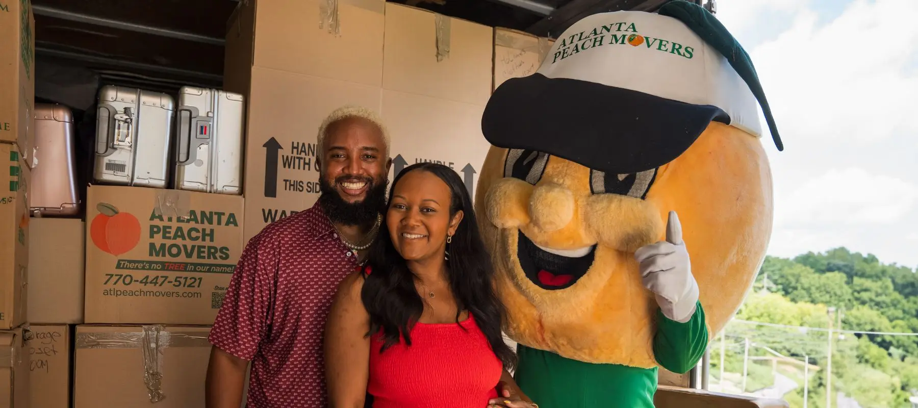 A smiling couple poses with a person in an Atlanta Peach Movers peach mascot costume, surrounded by stacked moving boxes outdoors.