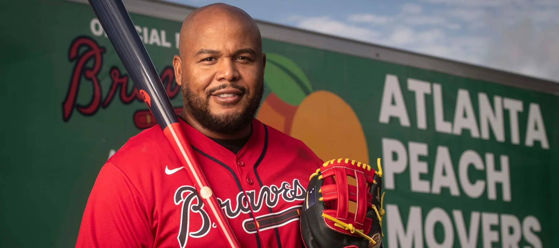 A man in a red Braves baseball jersey holds a bat and glove, standing in front of a sign that reads Atlanta Peach Movers with a peach graphic and blue sky in the background.