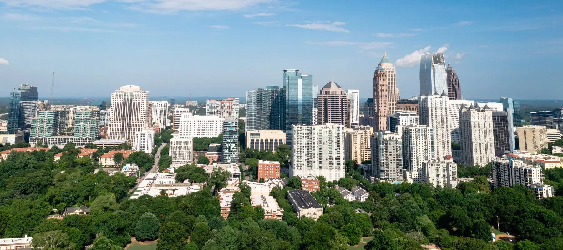 Aerial view of a city skyline with many modern skyscrapers and tall buildings, bordered by a large area of green trees and residential neighborhoods under a blue sky.