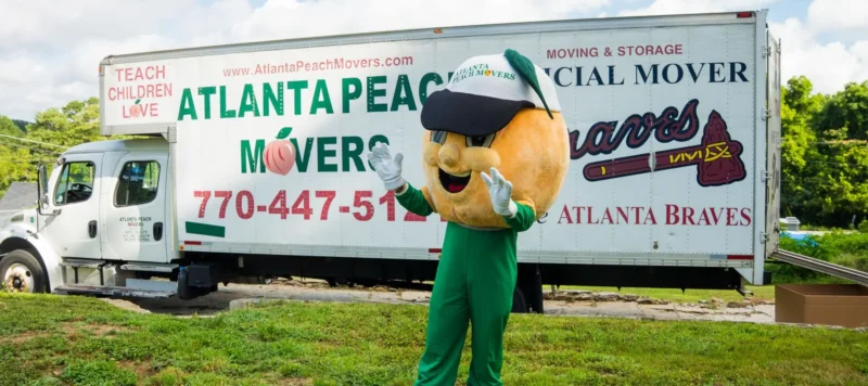 A person in a peach mascot costume waves in front of an Atlanta Peach Movers truck, which is parked on grass with trees in the background.