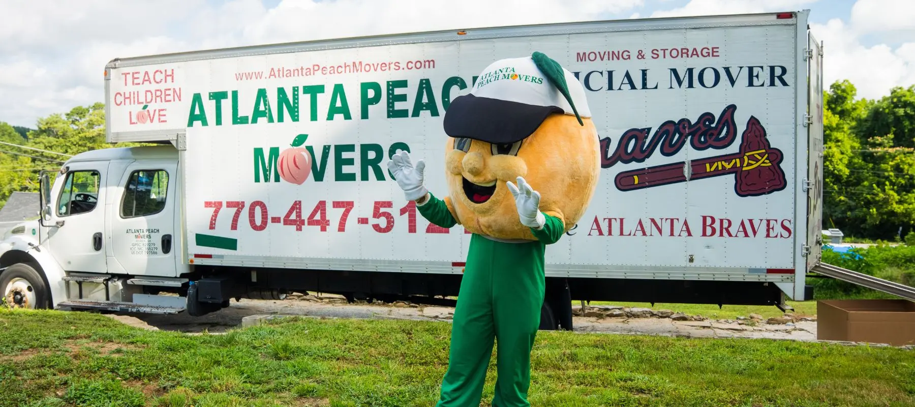 A person in a peach mascot costume waves in front of an Atlanta Peach Movers truck, which is parked on grass with trees in the background.