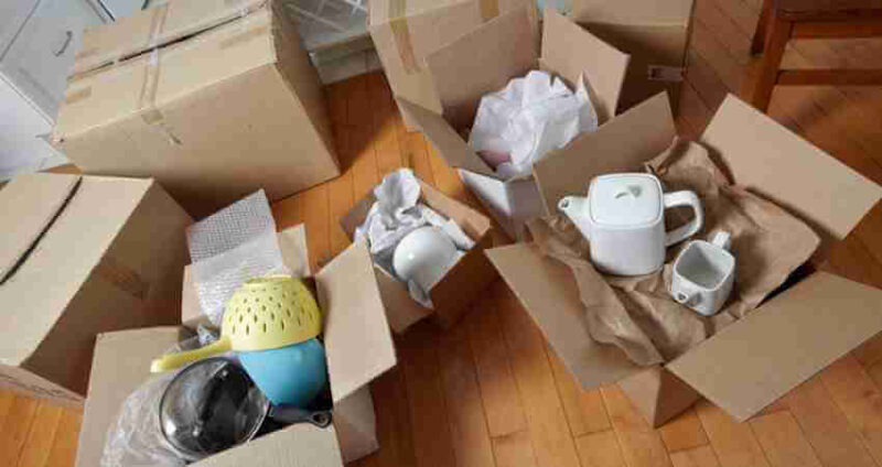 Several cardboard boxes on a wooden floor, some open and filled with kitchen items like bowls, a colander, a teapot, and cups, wrapped in paper for moving or packing.