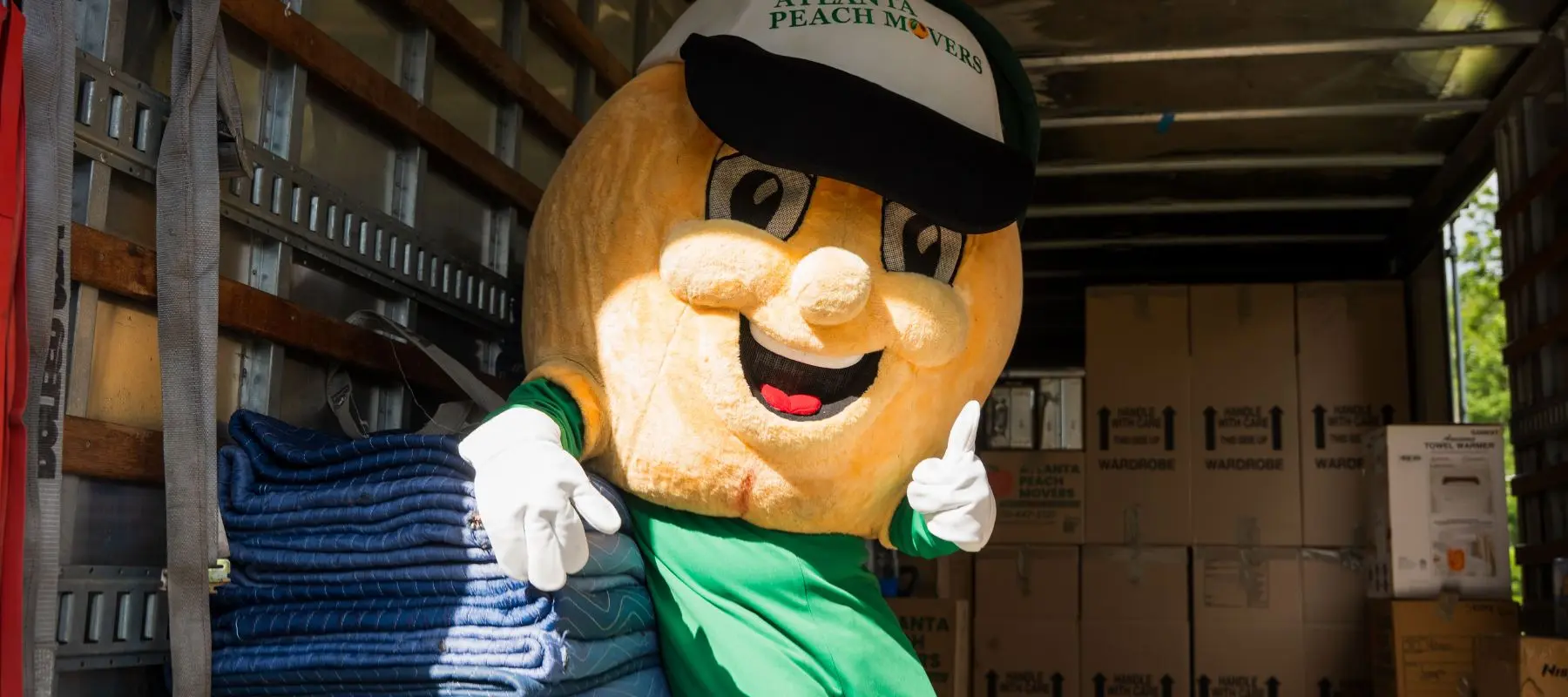 A person in a giant peach mascot costume stands in a moving truck filled with boxes, wearing a hat that says Atlanta Peach Movers, and gives a thumbs-up gesture with both hands.