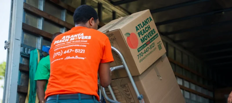 A mover in an orange Atlanta Peach Movers shirt loads a large box labeled Atlanta Peach Movers onto a truck using a dolly. Another person in green is partly visible nearby.