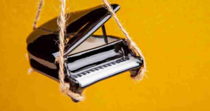 A small black grand piano model is suspended by ropes against a bright yellow background.