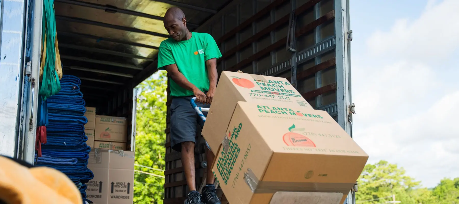 A person in a green shirt moves a large cardboard box with a dolly inside a truck. Several more boxes are stacked nearby, and trees are visible outside.