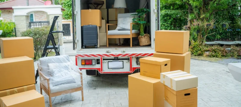 A moving truck is parked with its back open, showing packed furniture and boxes inside. Several cardboard boxes and wrapped furniture are on the driveway, ready to be loaded or unloaded.