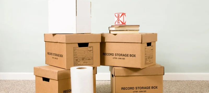 Five cardboard storage boxes are stacked on the floor, with a roll of bubble wrap, books, and a red fragile sign on top. A white box is on the stack. The background is a light-colored wall and carpeted floor.