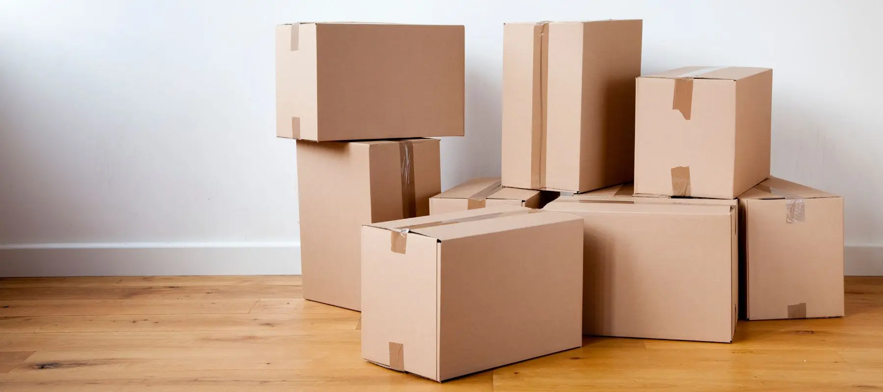 A group of sealed cardboard boxes stacked on a wooden floor against a white wall, ready for moving or storage.