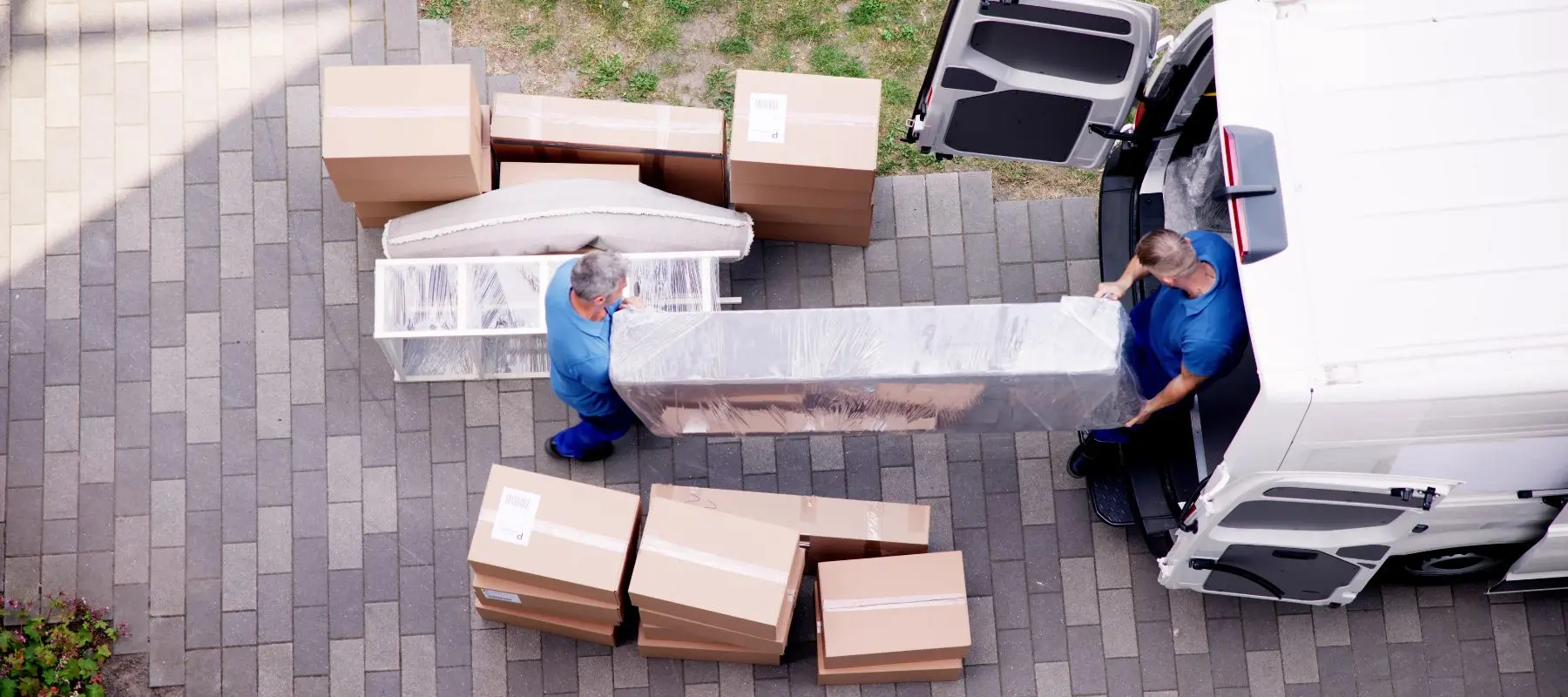 Two people in blue uniforms move a large wrapped object from a van onto a paved driveway, surrounded by cardboard boxes and other wrapped items, viewed from above.
