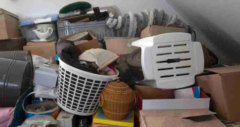 A cluttered attic space filled with boxes, a laundry basket, a white plastic chair, wicker baskets, and various household items stacked haphazardly under a slanted ceiling.