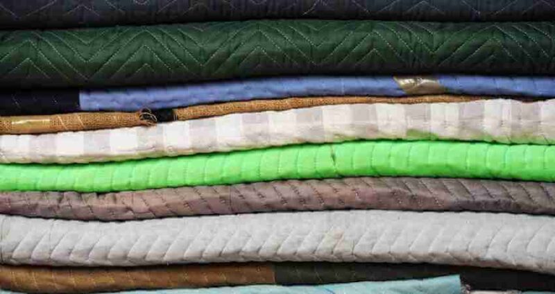 A close-up of several folded blankets and fabrics stacked on top of each other in various colors and textures, including green, gray, blue, and beige.