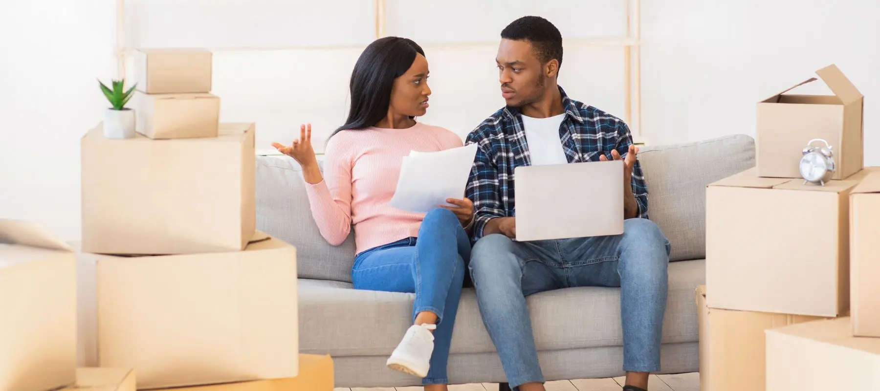 A couple sits on a couch surrounded by moving boxes, looking at each other with concerned expressions. One holds papers, the other a laptop, suggesting they are discussing moving or financial matters.
