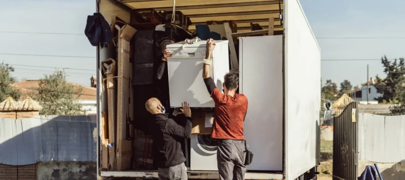 Two people load a truck with household items, including a washing machine and furniture, on a sunny day. The truck is parked outdoors near a fence and some buildings.