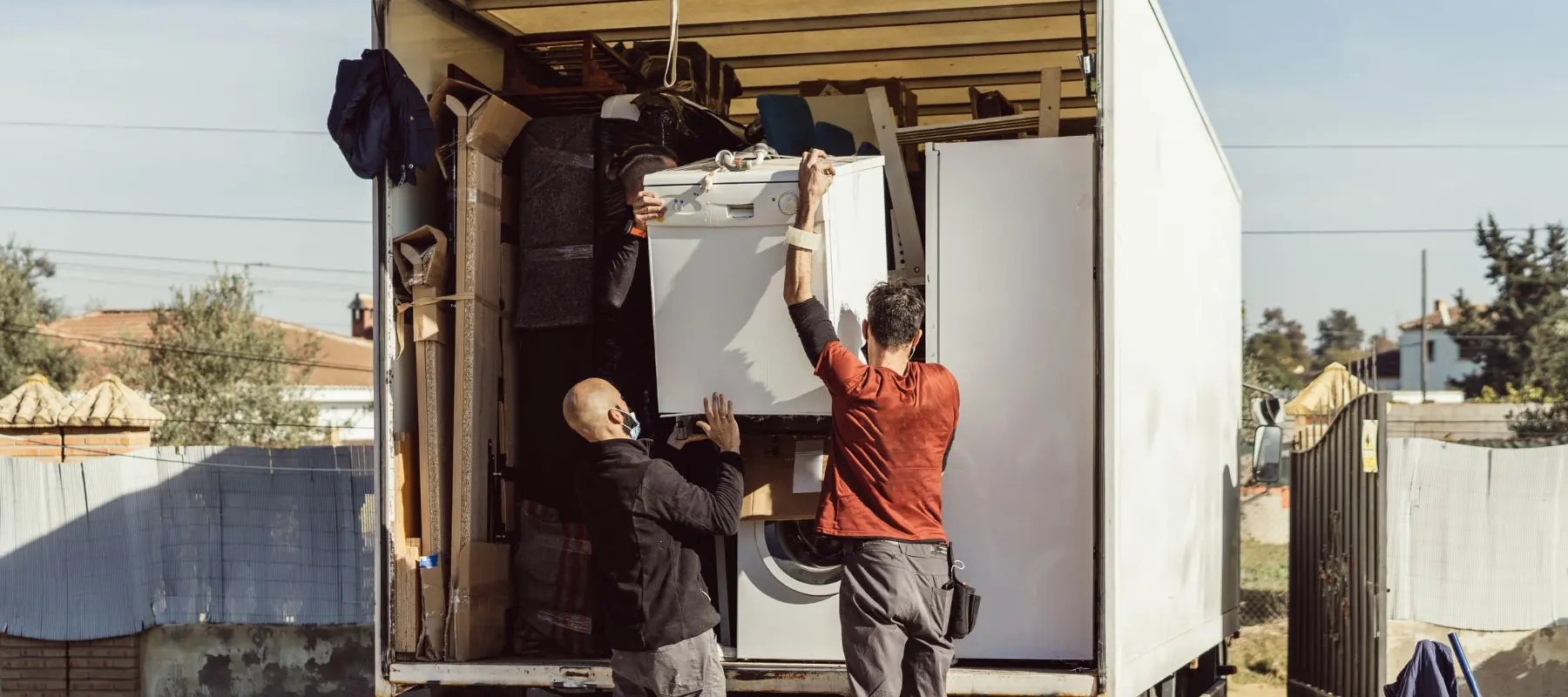 Two people load a truck with household items, including a washing machine and furniture, on a sunny day. The truck is parked outdoors near a fence and some buildings.