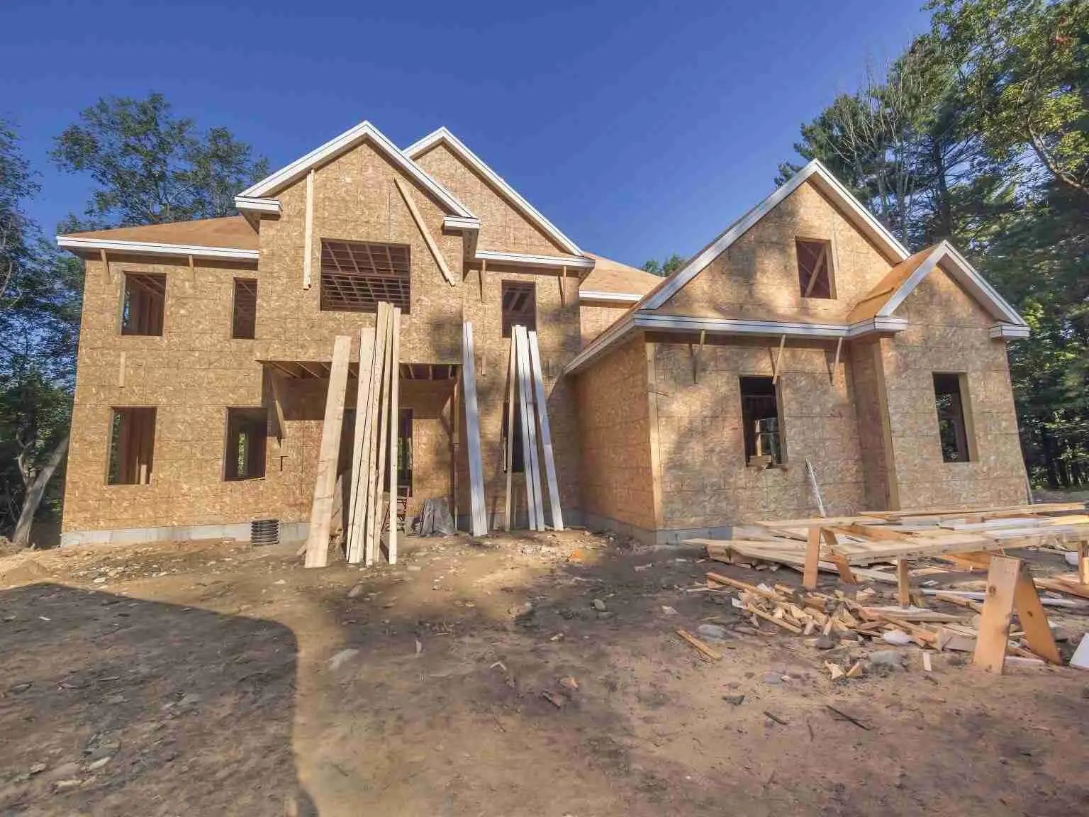 Partially constructed two-story house made of plywood, with exposed beams, open window frames, and building materials scattered on the ground under a clear blue sky.