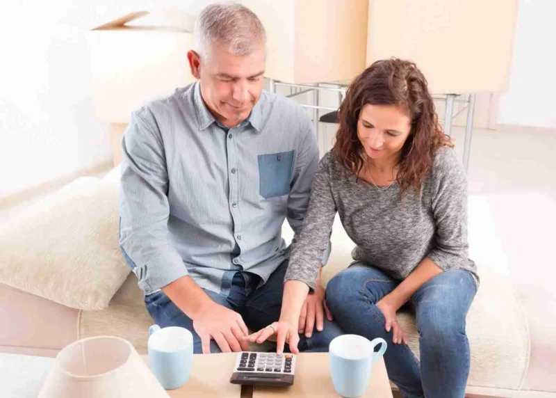 A man and woman sit on a couch, looking at a calculator on a table. Both have coffee mugs in front of them and appear to be discussing finances or budgeting in a bright living room.