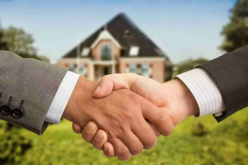 Two people in business suits shake hands outdoors with a large house in the background, suggesting a successful real estate deal or agreement.