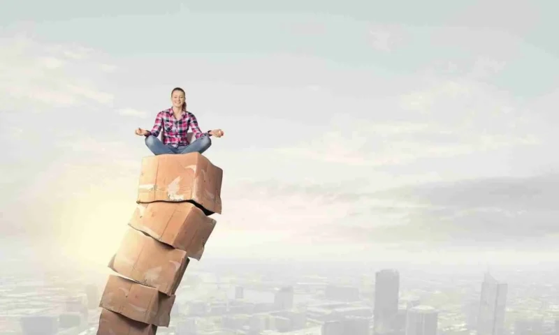 A person sits cross-legged in a meditative pose atop a tall, uneven stack of large stone blocks with a city skyline and cloudy sky in the background.