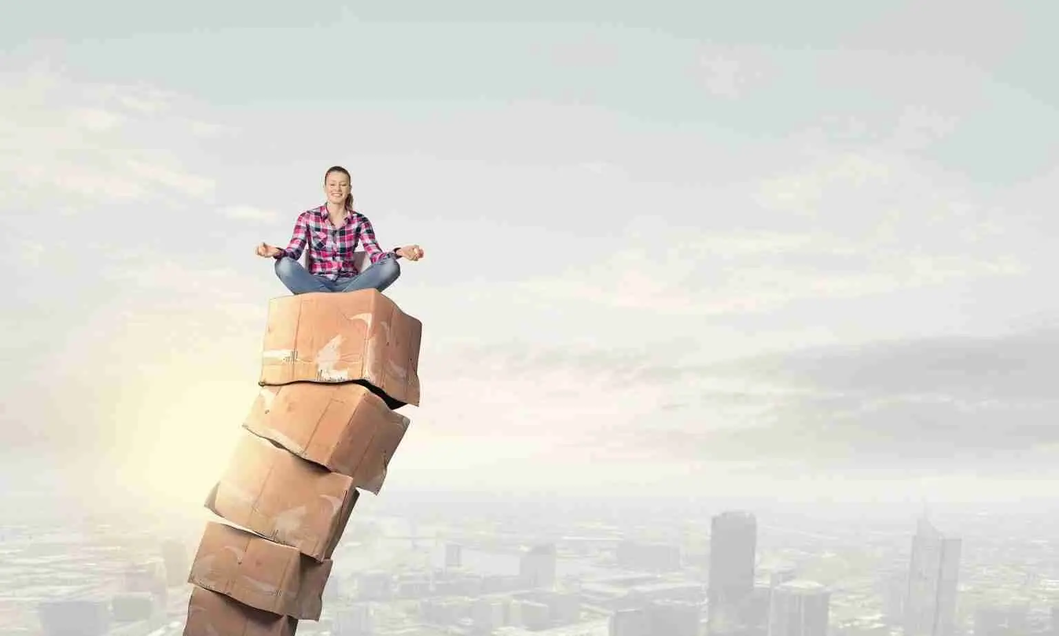 A person sits cross-legged in a meditative pose atop a tall, uneven stack of large stone blocks with a city skyline and cloudy sky in the background.