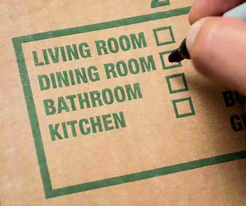 A hand marks the Dining Room checkbox on a cardboard moving box labeled with options for Living Room, Dining Room, Bathroom, and Kitchen.
