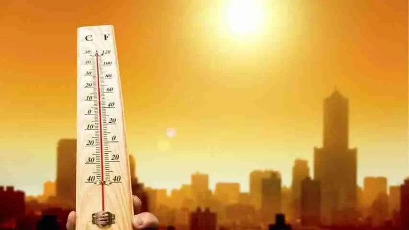 A person holds a thermometer showing high temperatures against a bright, orange sky with the sun shining over a city skyline, suggesting a hot day.