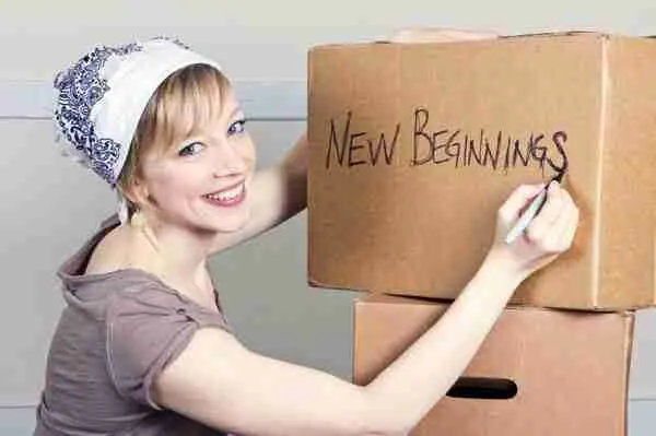 A smiling woman writes NEW BEGINNINGS on a cardboard box with a marker. She wears a light headscarf and casual clothes, sitting next to another box, suggesting moving or starting fresh.