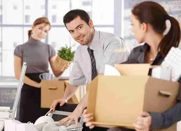 Three office workers pack up their belongings into boxes, preparing to move. One man smiles at a woman while organizing items, and another woman in the background holds a plant.