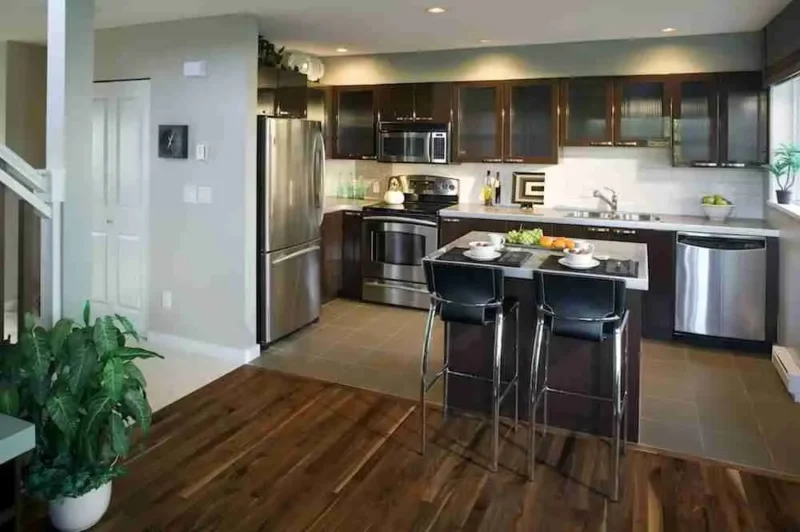 Modern kitchen with dark cabinets, stainless steel appliances, a kitchen island set for two, wooden and tile flooring, and a large green plant in the foreground. Ample natural light comes through a window above the sink.