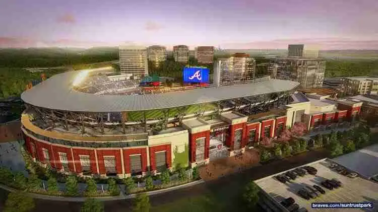 Aerial view of a modern baseball stadium with a large screen displaying the Atlanta Braves logo, surrounded by city buildings and parking areas at sunset.