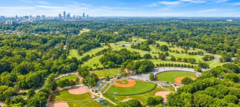 Aerial view of a large city park with baseball fields, a circular stadium, green lawns, many trees, and a distant city skyline under a blue sky with light clouds.