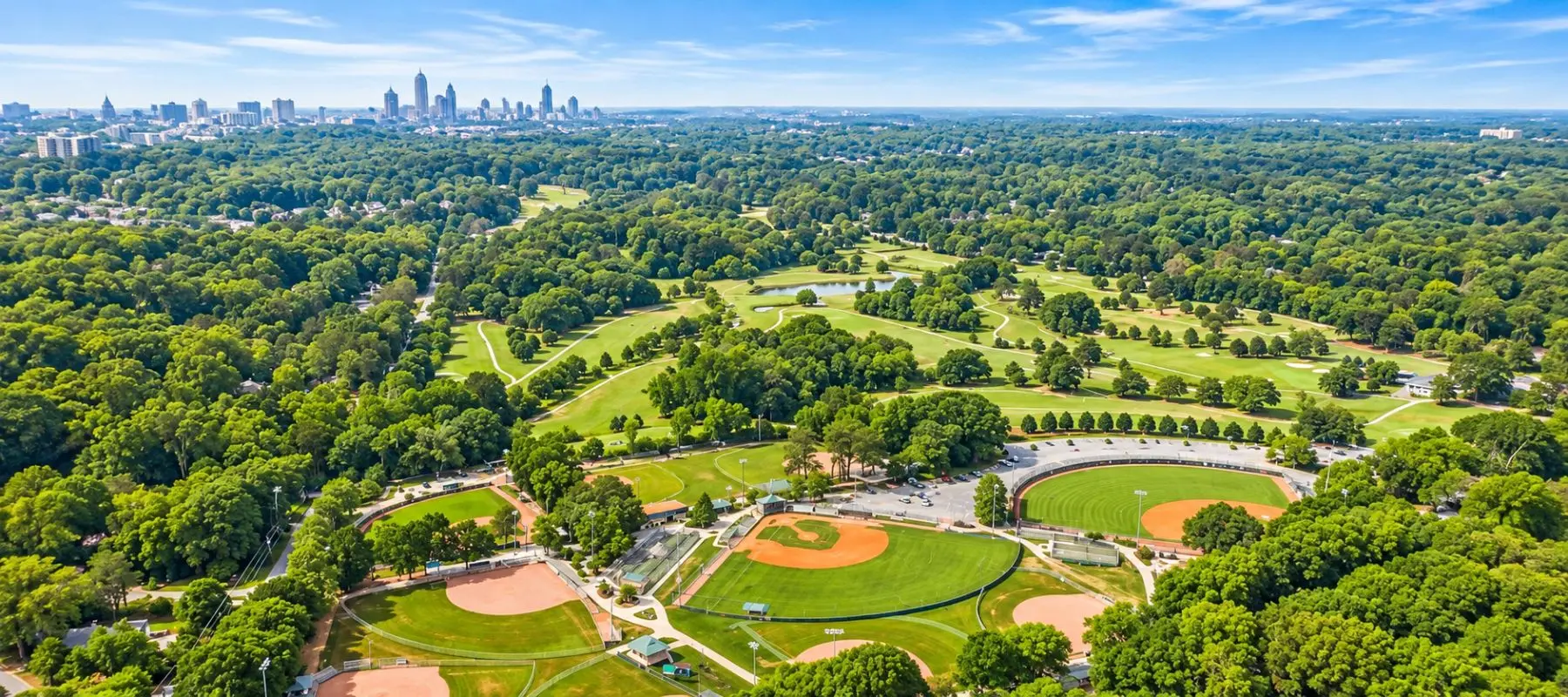 Aerial view of a large city park with baseball fields, a circular stadium, green lawns, many trees, and a distant city skyline under a blue sky with light clouds.