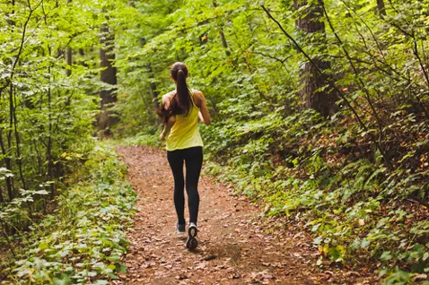 A person with long hair, wearing a yellow top and black leggings, jogs on a dirt path through a lush, green forest.