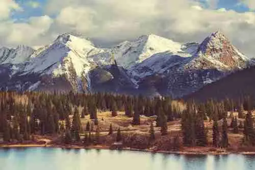 Snow-capped mountains rise behind a forest of evergreen trees, with a calm lake in the foreground under a partly cloudy sky.
