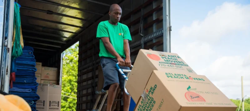 A man in a green shirt uses a dolly to move cardboard boxes labeled Atlanta Peach Movers inside a truck on a sunny day.