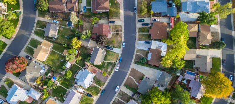 Aerial view of a suburban neighborhood showing houses, green lawns, trees, and cars parked along a curved street on a sunny day.