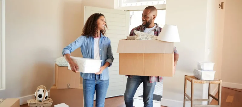 A smiling couple stands in a bright room holding moving boxes, surrounded by packed belongings, as they move into a new home.