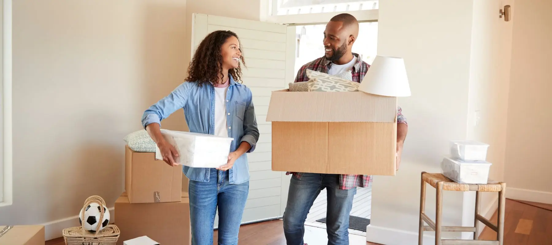A smiling couple stands in a bright room holding moving boxes, surrounded by packed belongings, as they move into a new home.