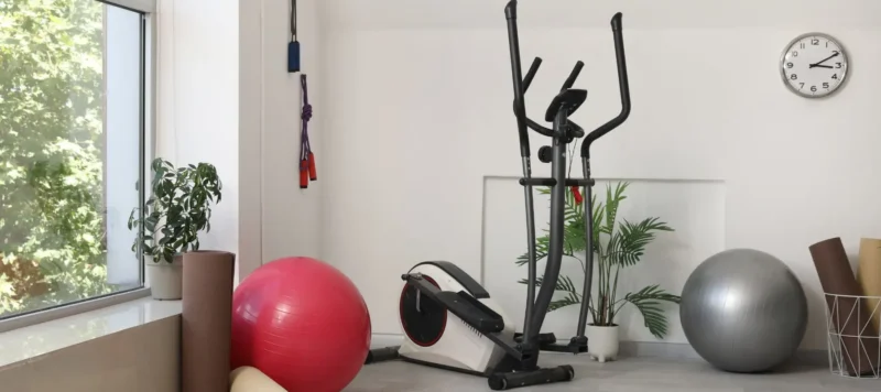 A home gym with an elliptical machine, a pink and a gray exercise ball, yoga mats, potted plants, jump ropes on the wall, and a clock showing 10:10. Sunlight streams through a window.