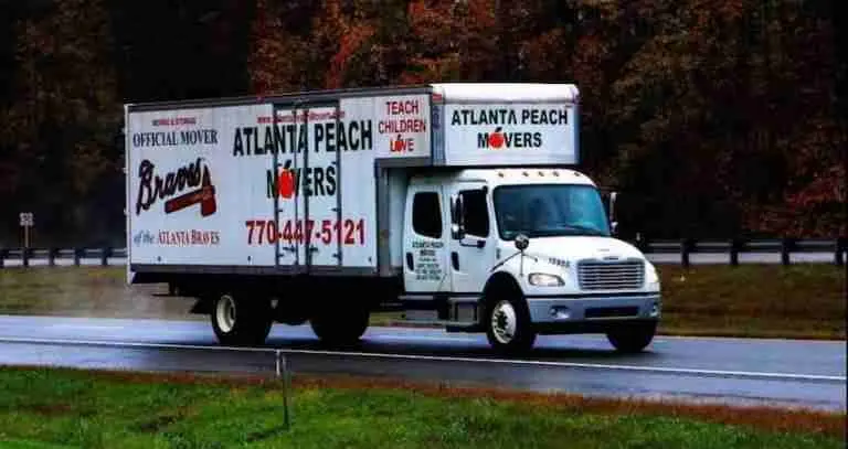 A white Atlanta Peach Movers truck drives on a wet road. The truck displays the Braves logo, phone number, and slogans including Teach Children Love and Official Movers. Trees with fall foliage are in the background.