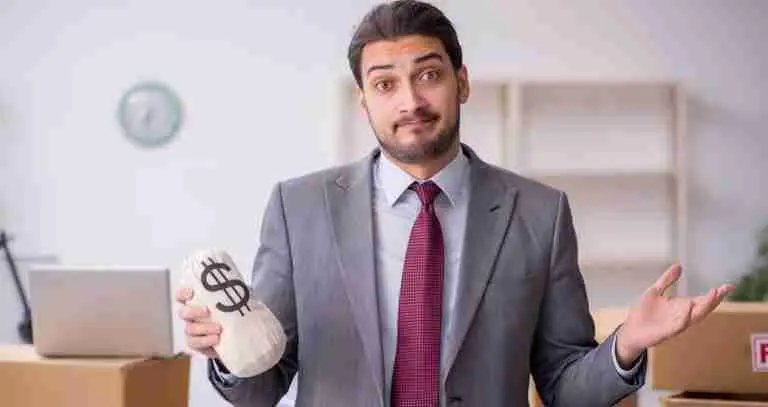 A man in a suit shrugs while holding a money bag with a dollar sign on it, standing in an office with cardboard boxes and a laptop in the background.