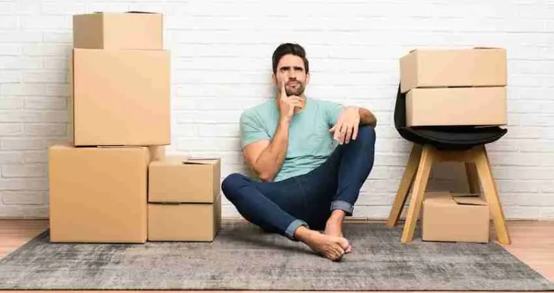 A man sits barefoot on the floor, surrounded by stacked cardboard moving boxes and a stool, looking thoughtful, with a white brick wall in the background.