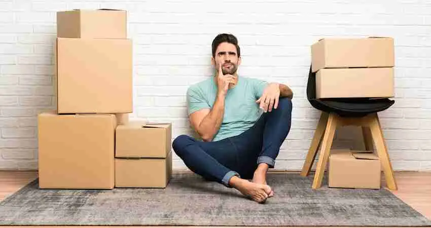 A man sits barefoot on the floor, surrounded by stacked cardboard moving boxes and a stool, looking thoughtful, with a white brick wall in the background.