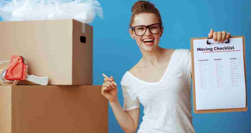 Smiling person wearing glasses holds a moving checklist on a clipboard, standing next to stacked cardboard boxes, packing tape, and bubble wrap, against a blue background.