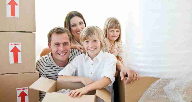 Smiling family with two kids surrounded by moving boxes. One child is sitting inside an open box, and the parents kneel behind them. The scene suggests they are moving into or out of a home.